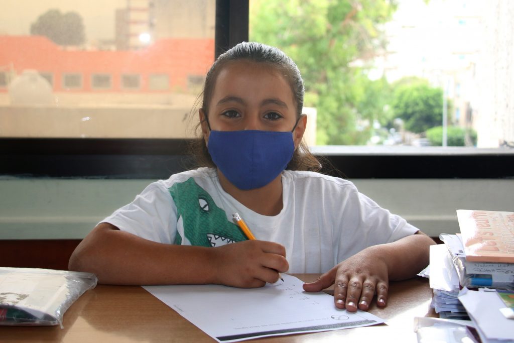 A Syrian refugee girl wearing a face mask sitting at a desk with paper and a pencil