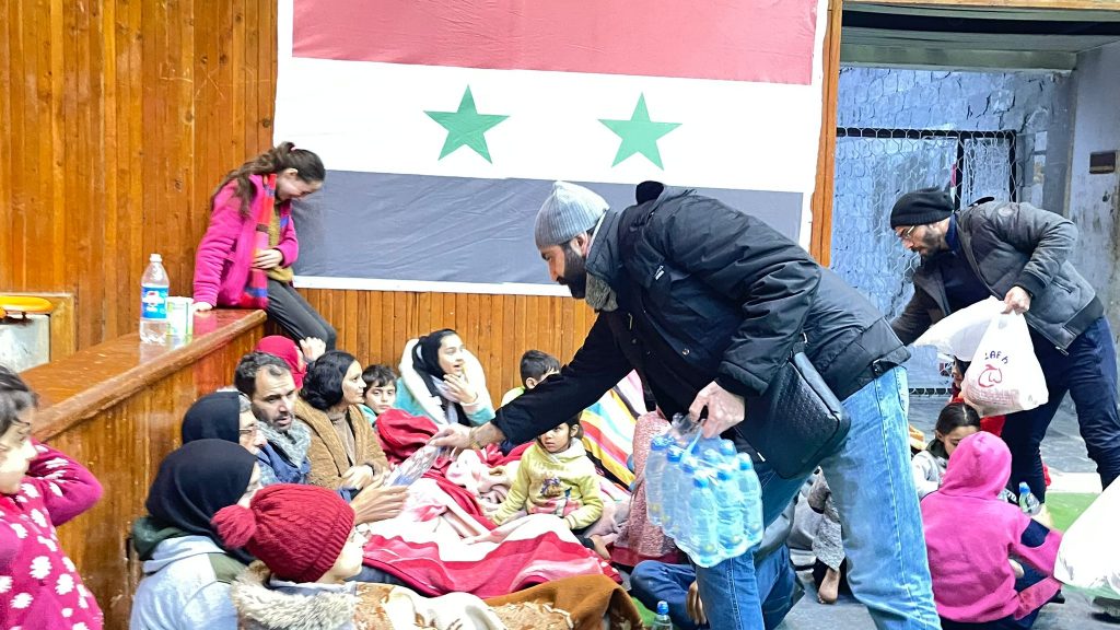 ADRA staff hand out food and water to a group of men, women and children as part of the Turkey response. The Syrian flag is hung on the wall behind them.