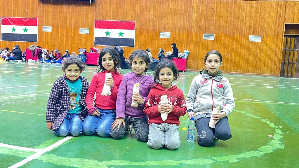 A group of young girls are eating wraps that were provided as part of ADRA's Turkey response. They are smiling at the camera and sitting in the middle of a basketball court in a school auditorium. Two Syrian flags are hung on the wall behind them.