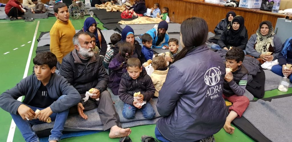 A group of men, women and children are sitting on mats eating wraps. They are facing a woman with an ADRA jacket who is helping coordinate the Turkey response.
