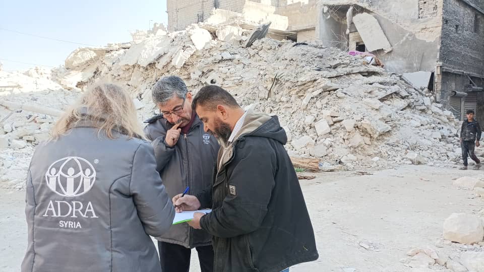 Three people standing in a huddle looking over documents with the rubble of a collapsed building in the background